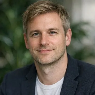 Man late 30s ash-blonde hair light eyes dark blazer white tee, indoor plant background